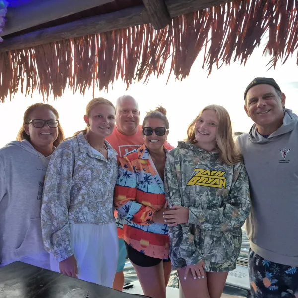 Six people smiling under a thatched roof near the water at sunset.