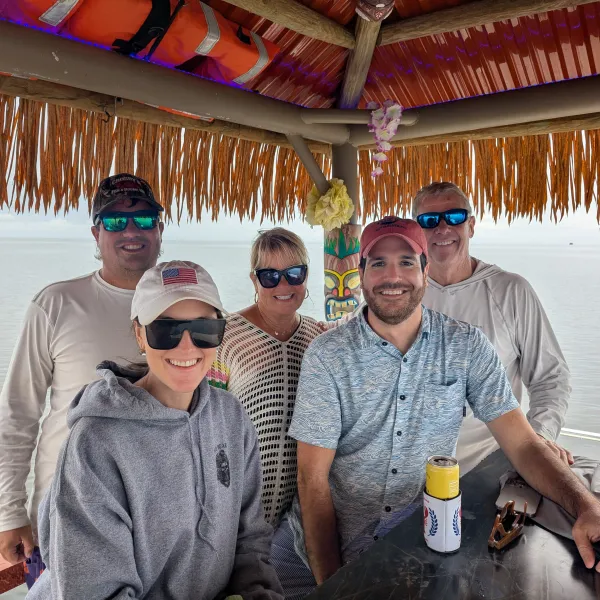 Five people smiling under a thatched roof by the water, wearing casual clothes and sunglasses.