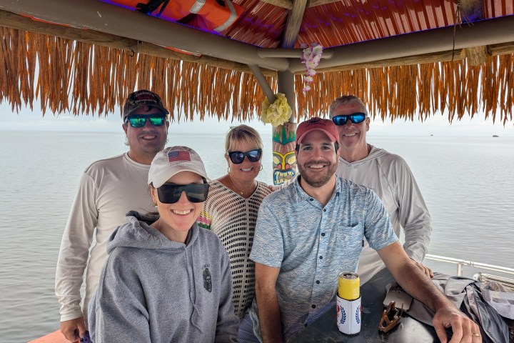 Five people smiling under a thatched roof by the water, wearing casual clothes and sunglasses.
