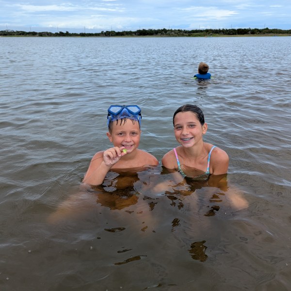 Two children smiling in shallow water, one with goggles, another person in the background.