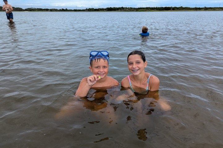 Two children smiling in shallow water, one with goggles, another person in the background.