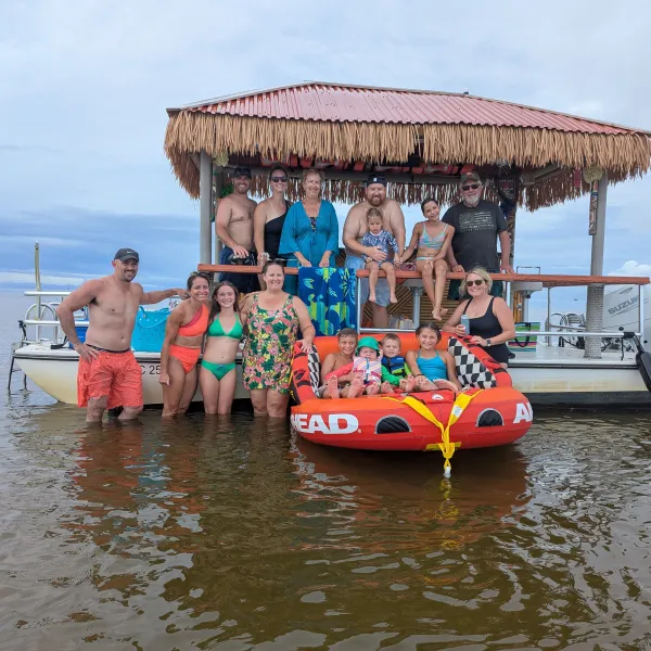 Group of people on a boat and tube in shallow water, under a thatched roof.