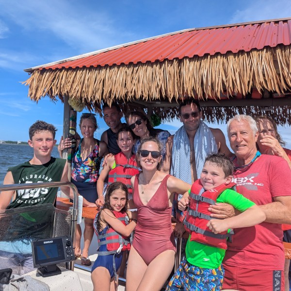 Group of people in swimwear on a boat with a thatched roof, smiling under a sunny blue sky.