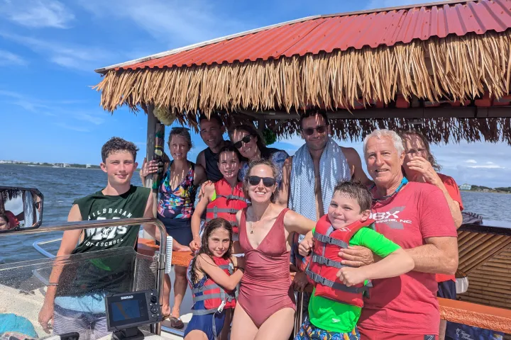 Group of people in swimwear on a boat with a thatched roof, smiling under a sunny blue sky.
