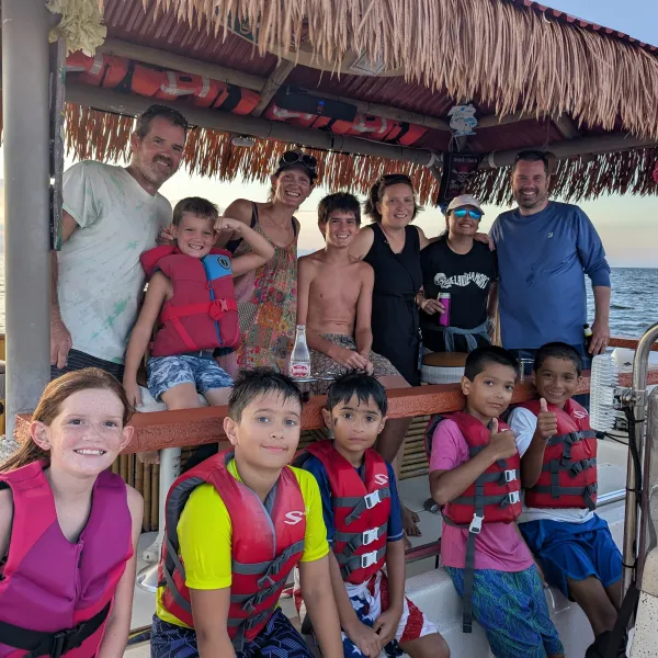Group of adults and kids wearing life jackets on a boat with tiki roof, ocean in the background.