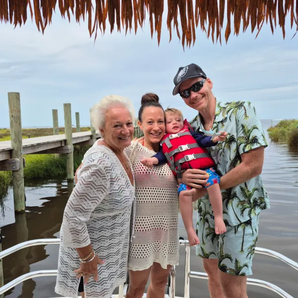 Four people, including a baby, smiling on a boat with a wooden dock and water in the background.