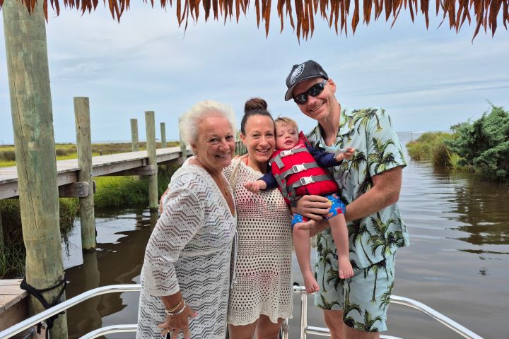 Four people, including a baby, smiling on a boat with a wooden dock and water in the background.