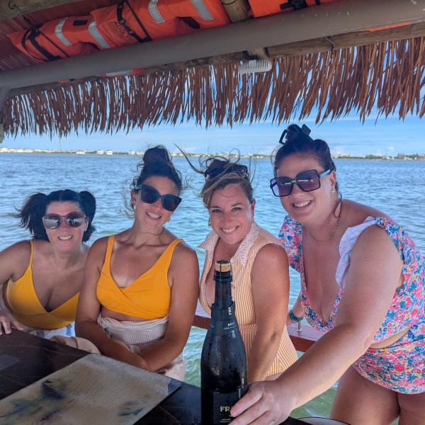 Four women in swimsuits under a tiki hut by the water, with one holding a bottle.
