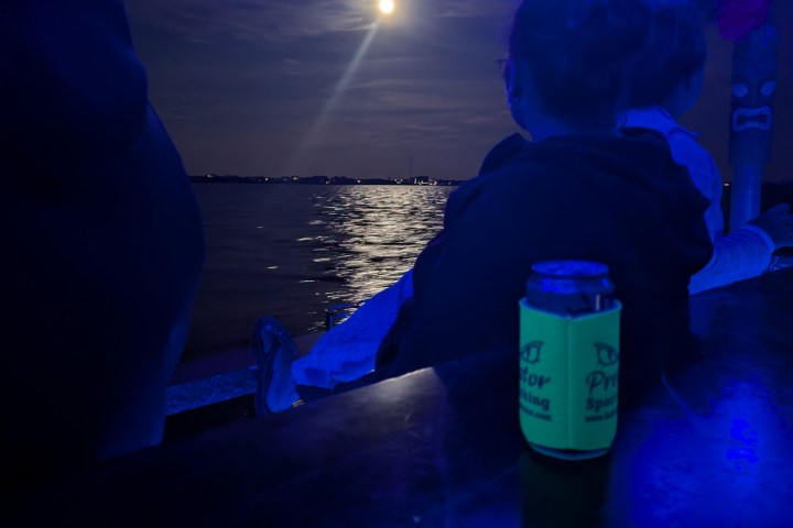 People sitting by water at night under blue lights, full moon in sky.